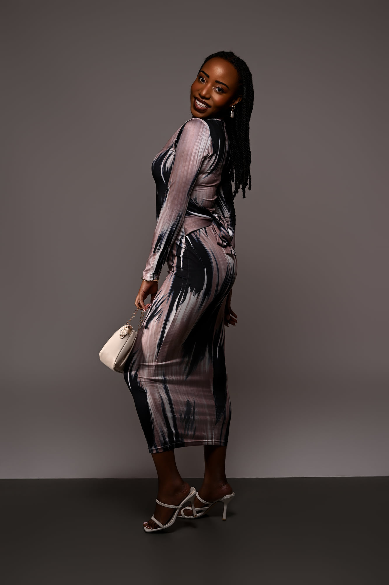 Lorna Ishimwe standing in a printed midi dress with a cream clutch, grey studio backdrop.