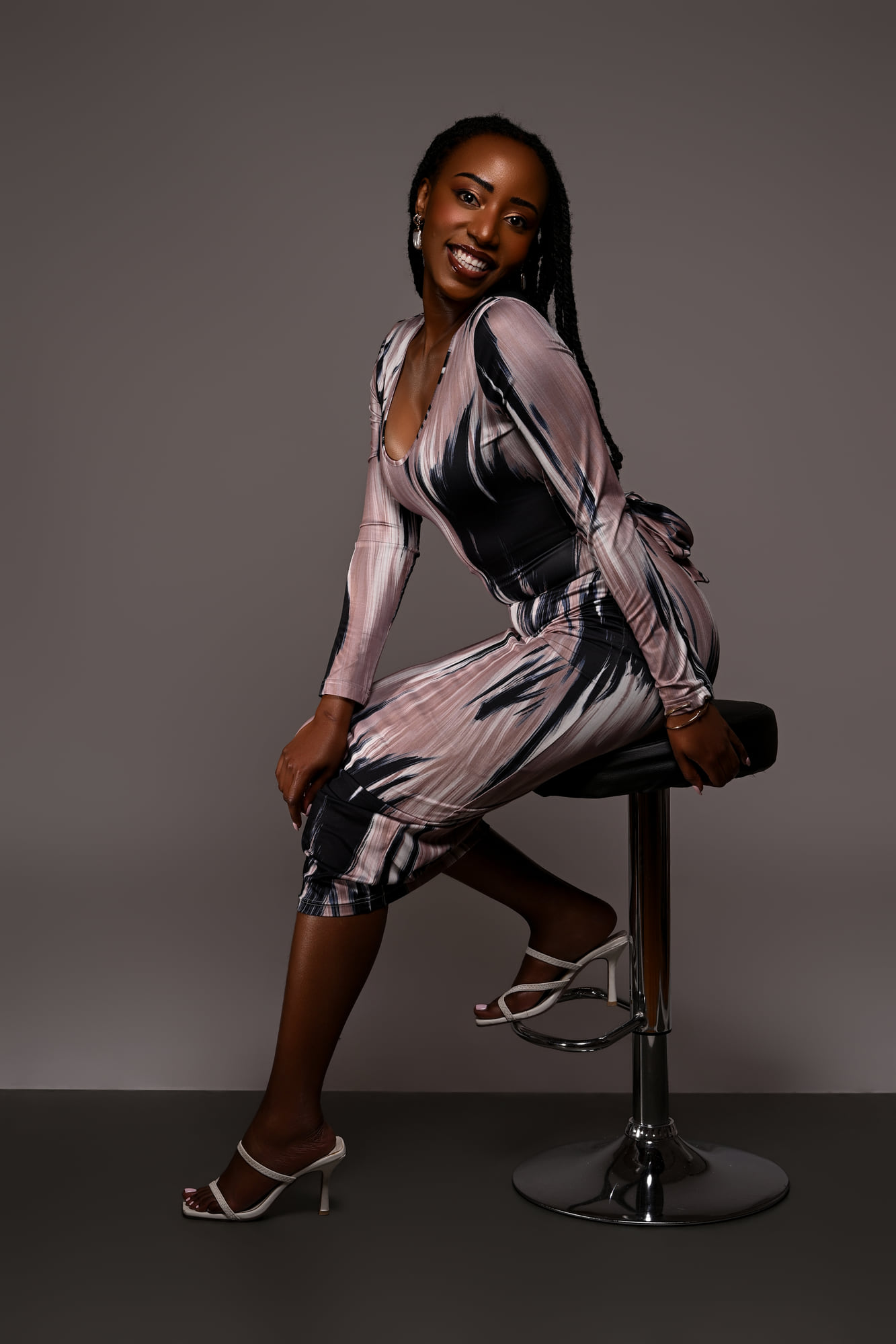 Lorna Ishimwe seated with one heel raised on a studio stool, printed midi dress.